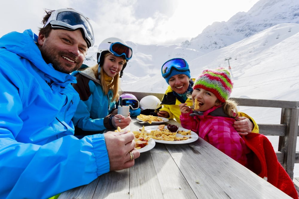Familie på skiferie der spiser mad med bjerge i baggrunden